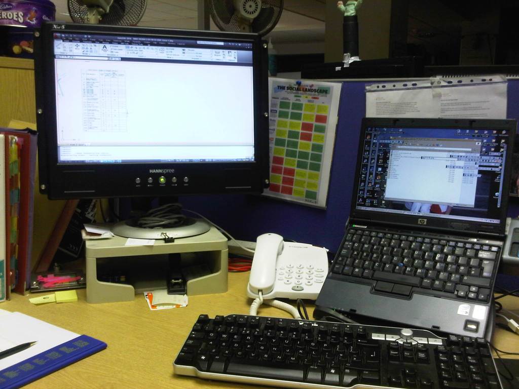 A typical cubical desk with a laptop, telephone and monitor. It is pretty messy.