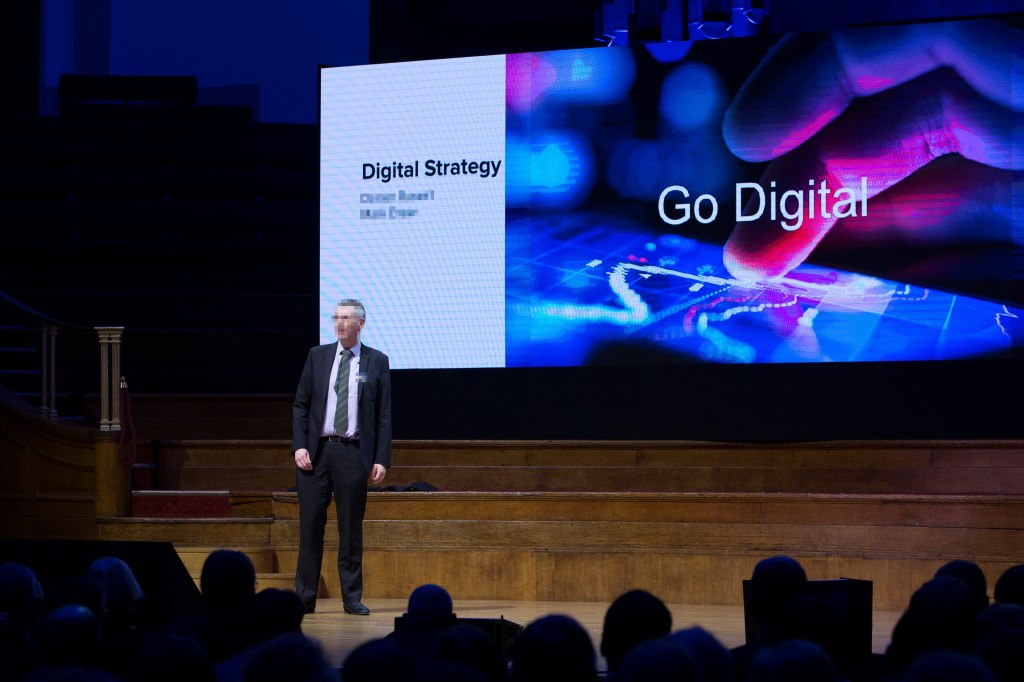 An executive addressing an audience. The screen behind him contains a message to "Go Digital"